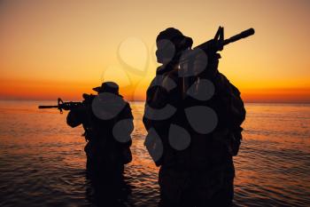 Two commando soldiers, army special operations fighters aiming assault rifles while coming out from water on shore at sunset or dawn. Coast guard shooters landing on sea coast, sneaking in water