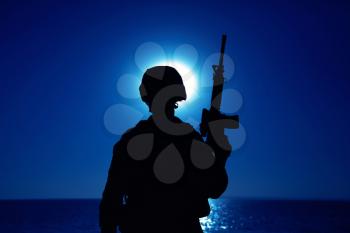 Silhouette of army soldier standing with service rifle in hand on background of night sky with moon. Armed Marines infantry in battle helmet and uniform ready for action, patrolling area in darkness