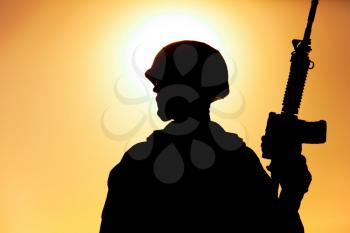 Silhouette of army soldier, special operations forces infantry rifleman, Marines fighter in combat helmet and ammunition standing with assault rifle in hand on background of sunset or dawn sky