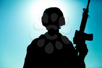 Silhouette of army soldier, special operations forces infantry rifleman, Marines fighter in combat helmet and ammunition standing with assault rifle in hand on background of sunset or dawn sky