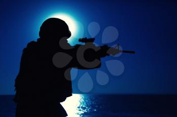 Silhouette of army special forces rifleman, soldier in ammunition and helmet aiming gun, shooting with service rifle on seacoast at night. Commando fighter, marine raider during amphibious mission