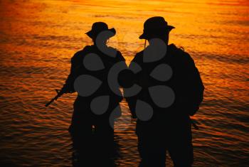 Two commando soldiers, army special operations fighters aiming assault rifles while coming out from water on shore at sunset or dawn. Coast guard shooters landing on sea coast, sneaking in water