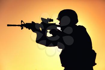 Silhouette of modern army infantry soldier standing on background of setting sun and aiming assault rifle with collimator sight. Special operations rifleman, elite forces shooter shooting in enemy