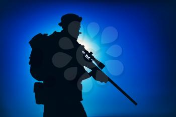 Silhouette of army elite forces fighter standing with sniper rifle on background of blue sky with moon or sun. Sniper or marksman in boonie hat, carrying backpack on mission, patrolling at night