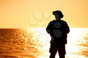Silhouette of army infantry soldier, coast guard fighter in boonie hat, standing on shore, sunset and ocean horizon on background. Border guard service soldier patrolling coastline, standing on beach