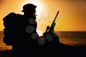 Silhouette of commando fighter, army special forces sniper sitting on sea or ocean shore on sunset. Coast guard rifleman observing beach, resting during coastline patrol or amphibious operation