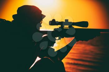 Commando team sniper, army special forces shooter aiming, shooting sniper rifle while sitting on sea or ocean shore during sunset. Coast or border guard soldier observing coastline with optical sight