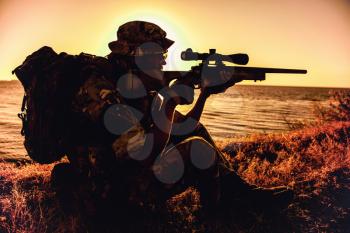 Commando team sniper, army special forces shooter aiming, shooting sniper rifle while sitting on sea or ocean shore during sunset. Coast or border guard soldier observing coastline with optical sight