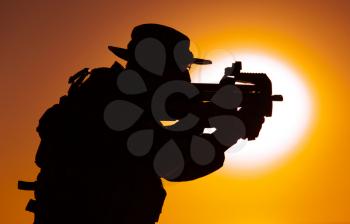 Silhouette of army special forces commando, soldier in boonie hat aiming and shooting bullpup submachine gun on background of setting sun. Private military company mercenary engaging enemy at sunset