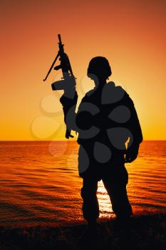Silhouette of army soldier, special operations shooter armed light machine gun standing on background of ocean or sea horizon at sunset time. Coast guard machine gunner on beach, patrolling coastline