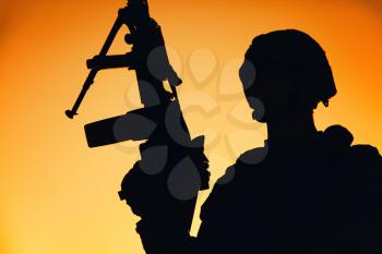 Shoulder silhouette of army machine gunner, special operations forces infantryman standing with raised gun on background of sunset sky. Marine Corps shooter in combat helmet holding weapon at dawn