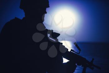 Silhouette of army infantryman standing with light machine gun on background of night sky with moon. Commando soldier in ammunition patrolling coastline, sneaking in darkness during night mission
