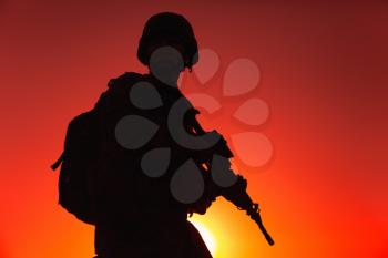 Silhouette of army rifleman in helmet and ammunition, carrying tactical backpack, walking with service rifle on background of sunset sky. Counter terrorist forces fighter marching at night mission