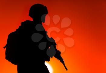 Silhouette of army rifleman in helmet and ammunition, carrying tactical backpack, walking with service rifle on background of sunset sky. Counter terrorist forces fighter marching at night mission