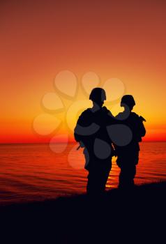 Silhouette of two Marines soldiers, special operations infantrymen or coast guard fighters in uniform and combat helmet, armed service rifles, standing on seashore, patrolling beach at sunset or dawn
