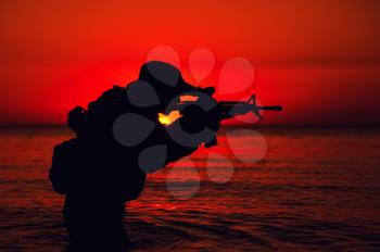 Silhouette of army soldier aiming and shooting service rifle while standing knee-deep in sea water. Coast guard, naval forces soldier attacking enemy with weapon, landing on beach on sunset or dawn