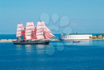 Sailing ship with red sails entering to the bay.
