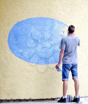 Young man paints blue oval on an external concrete wall. Adult male graffiti artist paints the wall. Urban outdoors street art concept. Place for text.
