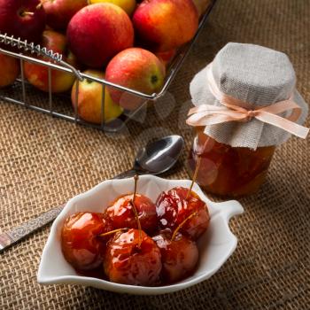 Cherry apples covered in syrup on white saucer, spoon, jar of homemade marmalade and apples in metal wire basket on a table covered with burlap, square. Selective focus. The toning