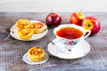 Cup of tea and small apple roses shaped pies. Sweet apple dessert pie. Homemade apple rose pastry. Breakfast tea with sweet apple pastry 