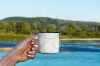 Woman holding a white campfire enamel mug mockup with summer river view. Empty mug mock up for design promotion.  