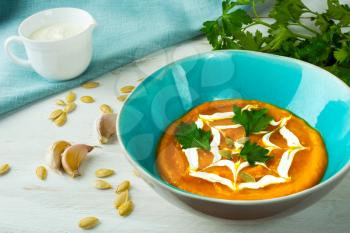 Pumpkin squash vegetable creamy soup topped with cream in a turquoise plate, pumpkin seeds, garlic and parsley on white wooden background 