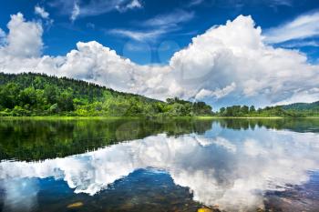 Beautiful landscape with cloudy blue sky reflected in the clear river water. Wooded waterside of a mountain lake. Summer idyllic landscape.