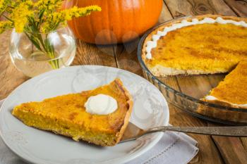Thanksgiving pumpkin pie slice on white plate, linen napkin, glass baking dish, vase, yellow flowers on a wooden background