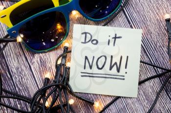 Do it now Conceptual handwritten message on white paper with wooden background and two sunglasses of different colors with lights in different order. Business concept handwritten messages.