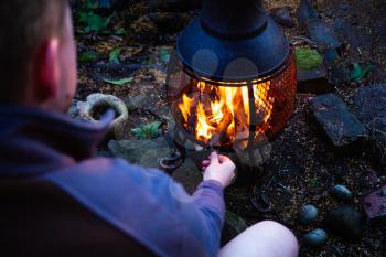 Camping concept with outdoor open fire flames. Camping fire and man in dark twilight moody photo. The man heats his hands in front of an open fire. Preparing barbecue fire outdoors.