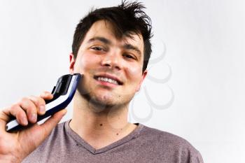 Smiling young man using electric shaver in front of mirror. Grooming and people concept. Handsome man using electric razor. Close up portrait of young handsome man with perfect skin and hair.