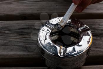 Burning cigarette in a metal ashtray. Healthcare concept with smelly and dirty cigarette. Hand holds burning cigarette above ashtrey. Lung cancer warning for smokers.