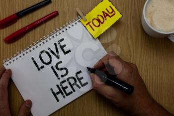 Handwriting text writing Love Is Here. Concept meaning Romantic feeling Lovely emotion Positive Expression Care Joy Man holding marker notebook clothespin reminder wooden table cup coffee