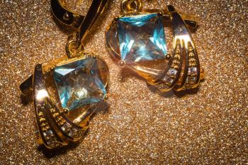 Modern geometric gold earrings decorated with light blue zirconia on glitter background.