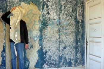 Male figure behind torn wallpaper shred in abandoned building interior.