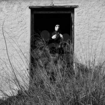 Masked figure by broken door of abandoned house obscured by overgrown plants.