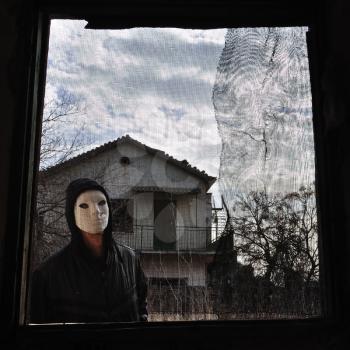 Hooded figure behind threaded window of derelict house. Masked maniac looking at you the viewer!