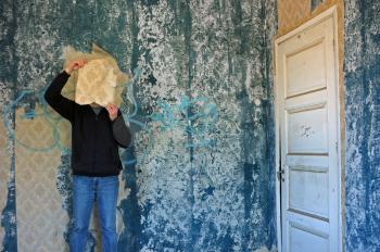 Man with torn wallpaper shred in decayed blue room abandoned house interior.
