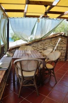 Wooden table and chairs, veranda with drapes and sea view. Mediterranean architecture.