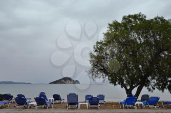 Lounge sunbed chairs with sea view tamarix tree and distant islet, Keri beach, Zakynthos Greece.