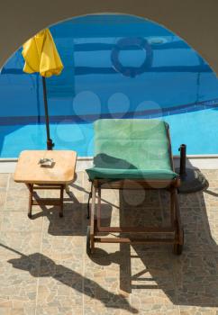 Green sunbathing chair with wooden table with yellow parasol in front of a blue pool
