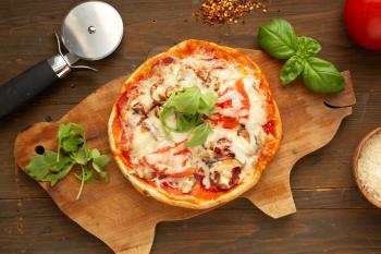 Homemade pizza with tomato, mushrooms, peppers and fresh herbs basil and arugula presented on a wooden board with pizza cutter