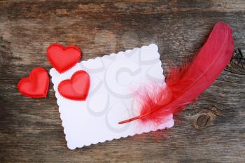 Blank white paper with red feather and satin hearts on it.  Wooden background.