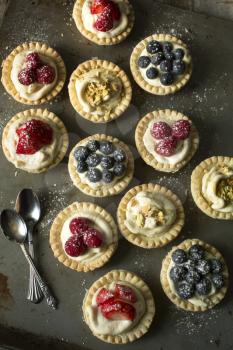 Blueberry, strawberry and pistachios tartlets with spoons on biscuit sheet on a wooden table