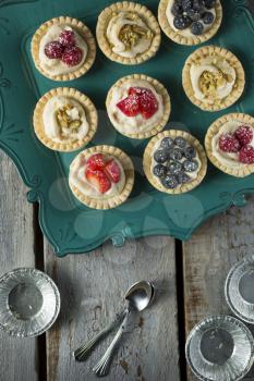 Blueberry, strawberry and pistachios tarts and spoons and empty aluminum plate in a green tray on a wooden table