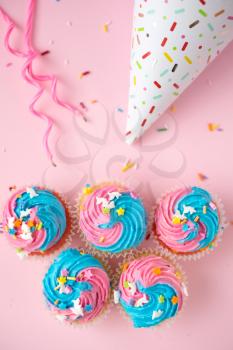 Top view of cupcakes with blue and pink icing, candles and birthday hat on a pink background