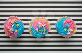 Three cupcakes with blue and pink icing on a black and white lined background