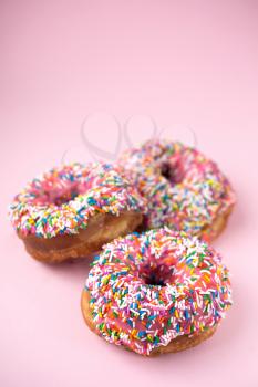 Three donuts with pink icing and candies on a pink pastel background