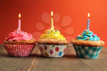 Vanilla cupcake in a row with yellow, pink and blue frosting and a birthday candle on a wooden table with a orange background