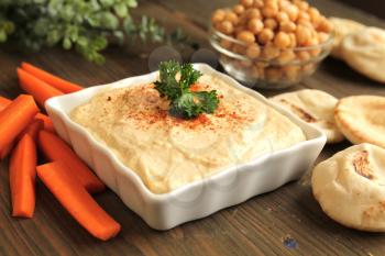 Bowl of chickpeas hummus with fresh parsley on top with pita bread and carrots to dip